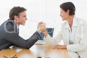 Smiling business couple arm wrestling at office desk