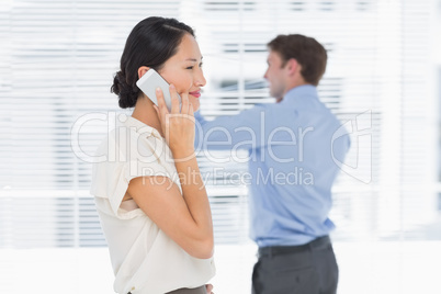 Businesswoman using cellphone with man behind at office