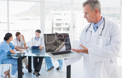 Doctor holding laptop with group around table in hospital