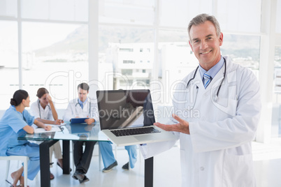 Doctor holding laptop with group around table in hospital