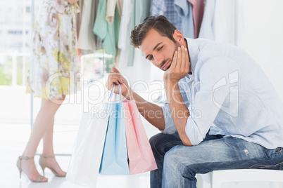 Bored man with shopping bags while woman by clothes rack