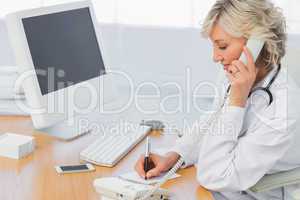 Female doctor using phone while writing notes at office