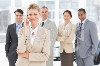 Businesswoman on the phone smiling at camera with team behind he Businesswoman on the phone smiling at camera with team behind he