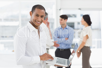 Young businessman using laptop with team behind him smiling at c