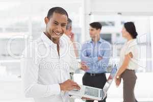 Young businessman using laptop with team behind him smiling at c