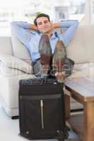 Smiling businessman sitting on couch with feet up on suitcase