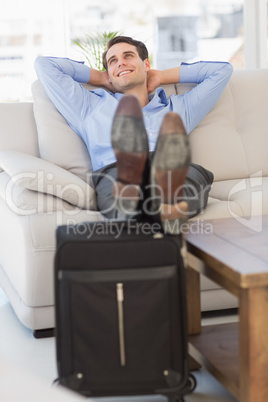 Smiling businessman relaxing on couch with feet up on suitcase