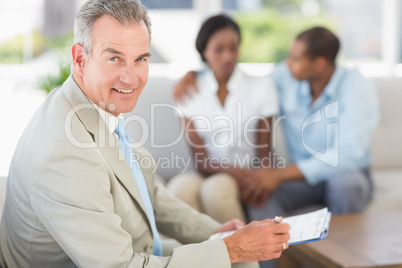 Salesman smiling at camera with couple on sofa behind him