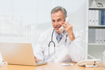 Smiling doctor using laptop and talking on phone at desk