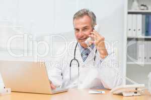 Smiling doctor using laptop and talking on phone at desk