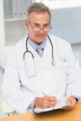 Smiling doctor sitting at his desk writing on clipboard
