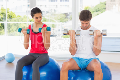 Couple lifting dumbbells while on fitness balls in gym