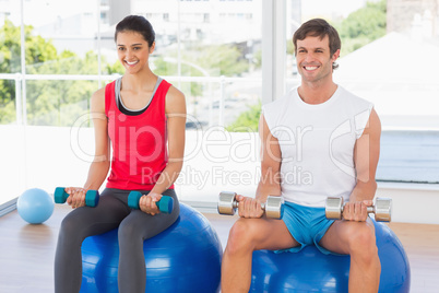 Couple lifting dumbbells while sitting on fitness balls in gym