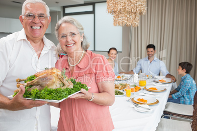 Grandparents holding chicken roast with family at dining table