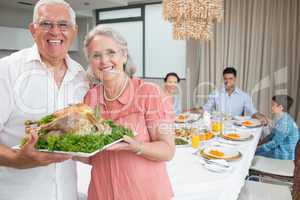 Grandparents holding chicken roast with family at dining table