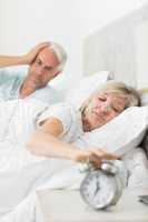 Man covering ears while woman extending hand to alarm clock in b