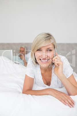 Happy woman using cellphone while man using laptop in bed
