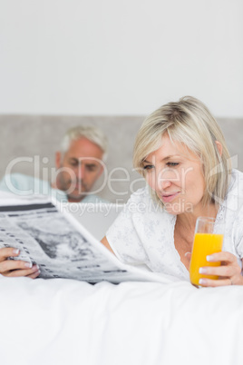 Couple reading newspaper and using laptop in bed