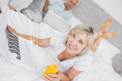 Couple reading newspaper and using laptop in bed