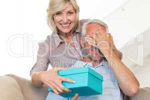 Smiling woman surprising mature man with a gift on sofa