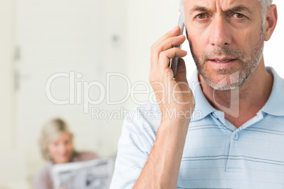 Mature man using cellphone with woman reading newspaper in backg