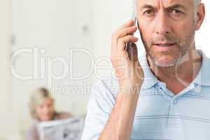 Mature man using cellphone with woman reading newspaper in backg