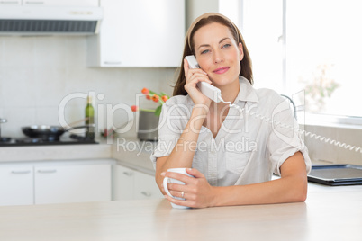 Smiling woman with coffee cup using landline phone in kitchen