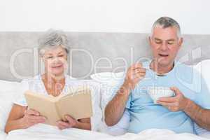 Senior couple with book and cereal bowl in bed