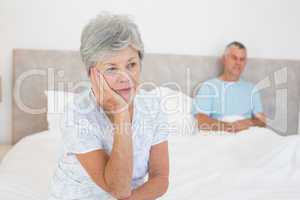 Sad senior woman on bed with husband in background