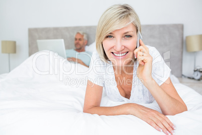 Happy woman using cellphone while man using laptop in bed