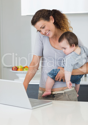 Mother using laptop while carrying baby in kitchen