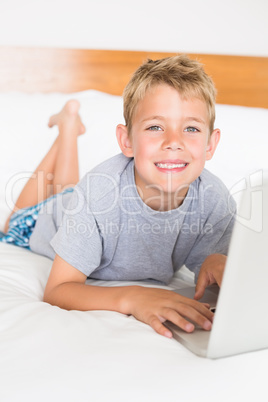 Blonde boy lying on bed using laptop smiling at camera