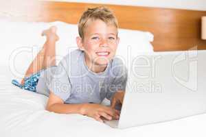 Happy blonde boy lying on bed using laptop