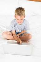 Cheerful blonde boy sitting on bed using laptop