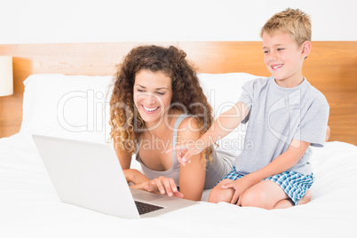 Cute blonde boy and mother on bed using laptop
