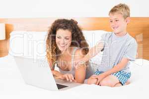 Cute blonde boy and mother on bed using laptop