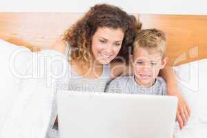 Cheerful mother and son sitting on bed using laptop