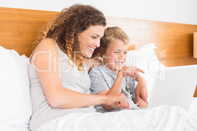 Smiling mother and son sitting on bed looking at laptop