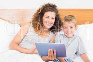 Smiling mother and son sitting on bed with tablet pc