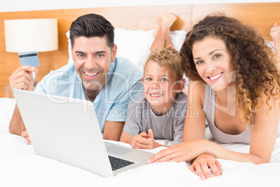 Smiling young family using laptop to shop online together on bed