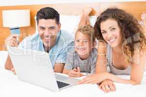 Smiling young family using laptop to shop online together on bed