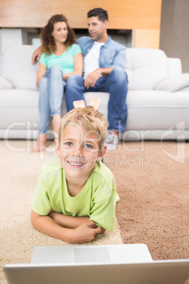 Happy little boy using laptop on the rug with parents sitting so