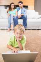 Cute little boy using laptop on the rug with parents sitting sof