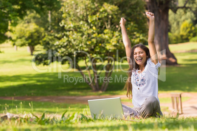 Cheerful woman raising hands with laptop in park