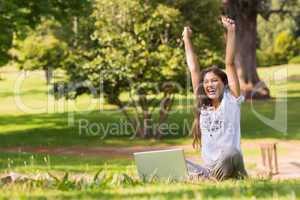 Cheerful woman raising hands with laptop in park