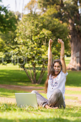 Cheerful woman raising hands with laptop in park