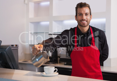 Smiling barista pouring milk into cup of coffee looking at camer