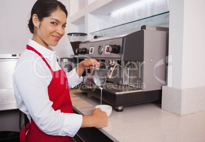 Pretty young barista pouring milk into cup of coffee smiling at