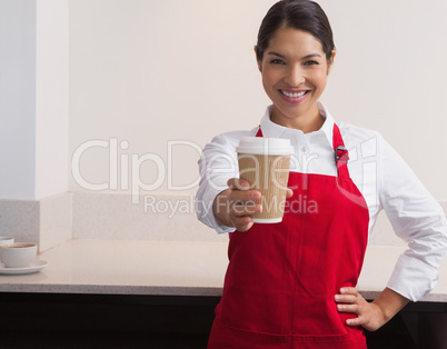 Pretty young barista offering cup of coffee to go smiling at cam