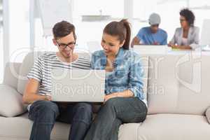 Couple using laptop with colleagues in background at creative of